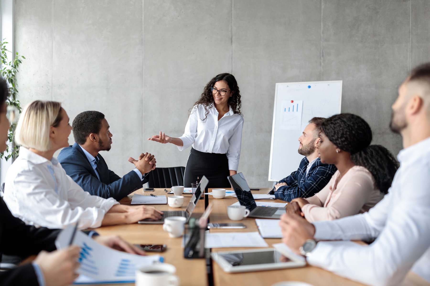 Eine Gruppe diverser junger Büromitarbeiter sitzt an einem Tisch und hört gut gelaunt einer weiblichen Führungskraft zu, die am Kopf des Tisches steht und etwas auf einem Whiteboard erklärt.