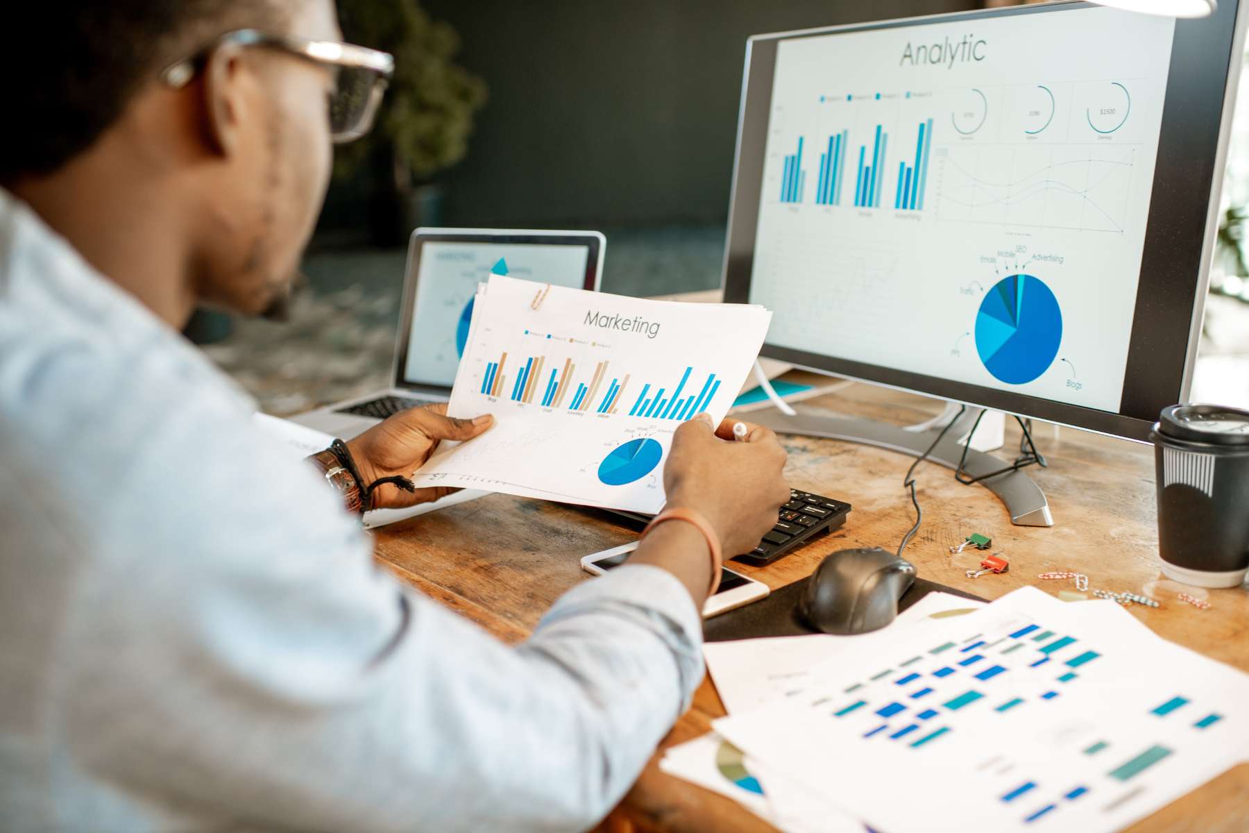 Ein junger Büroangestellter sitzt an einem Schreibtisch vor einem Bildschirm, auf dem Statistiken angezeigt werden und hält ein Blatt in der Hand, auf dem ebenfalls eine Statistik abgebildet ist.