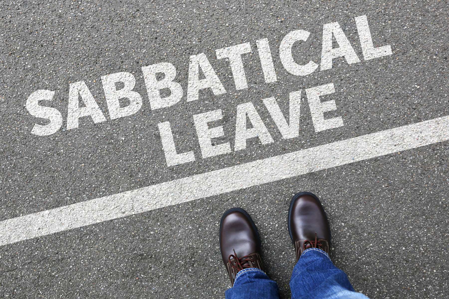 Aufnahme der Hosenbeine und Schuhe eines Mannes aus der Vogelperspektive, der auf der Straße vor einer weißen Linie steht, über welcher die Worte Sabbatical Leave stehen.