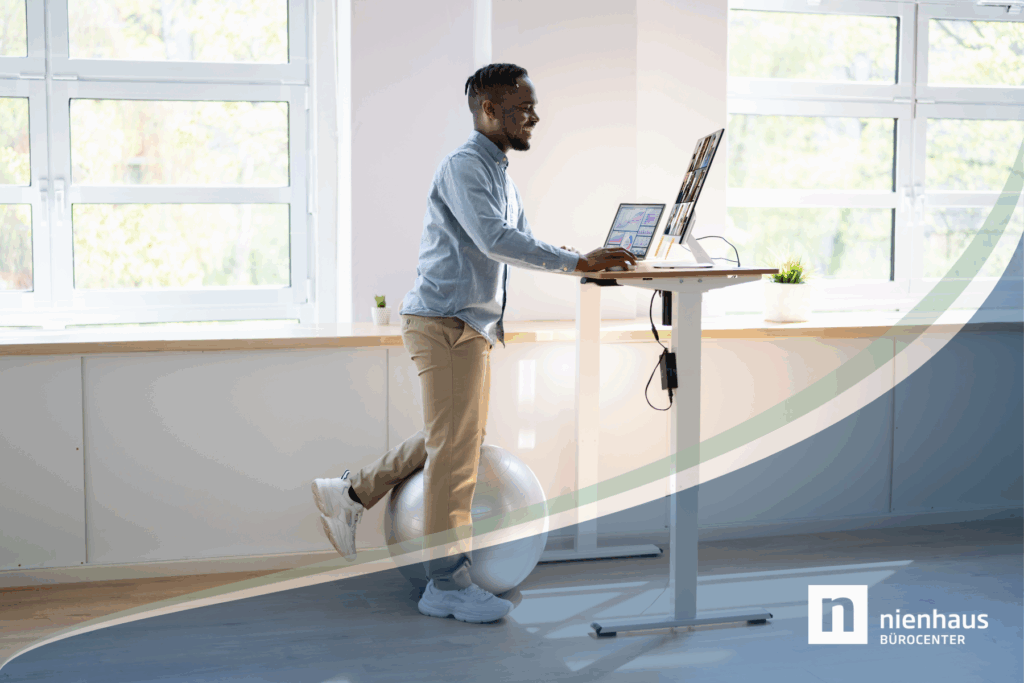 Junger Mann arbeitet im Büro an einem höhenverstellbaren Schreibtisch und stütz dabei ein Bein auf einen Gymnastikball.