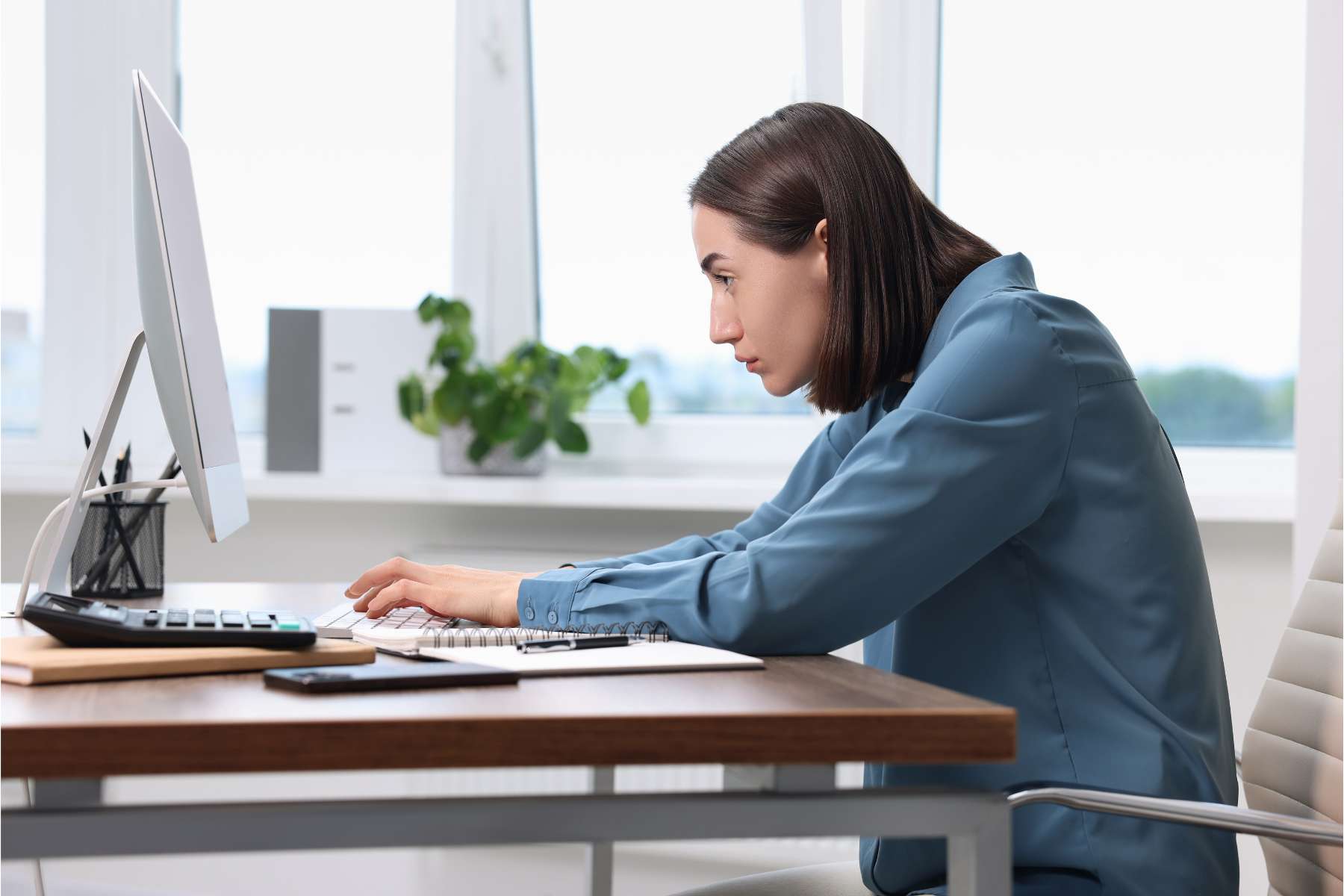 Junge Frau sitzt mit krummem Rücken am Schreibtisch und arbeitet am Laptop.