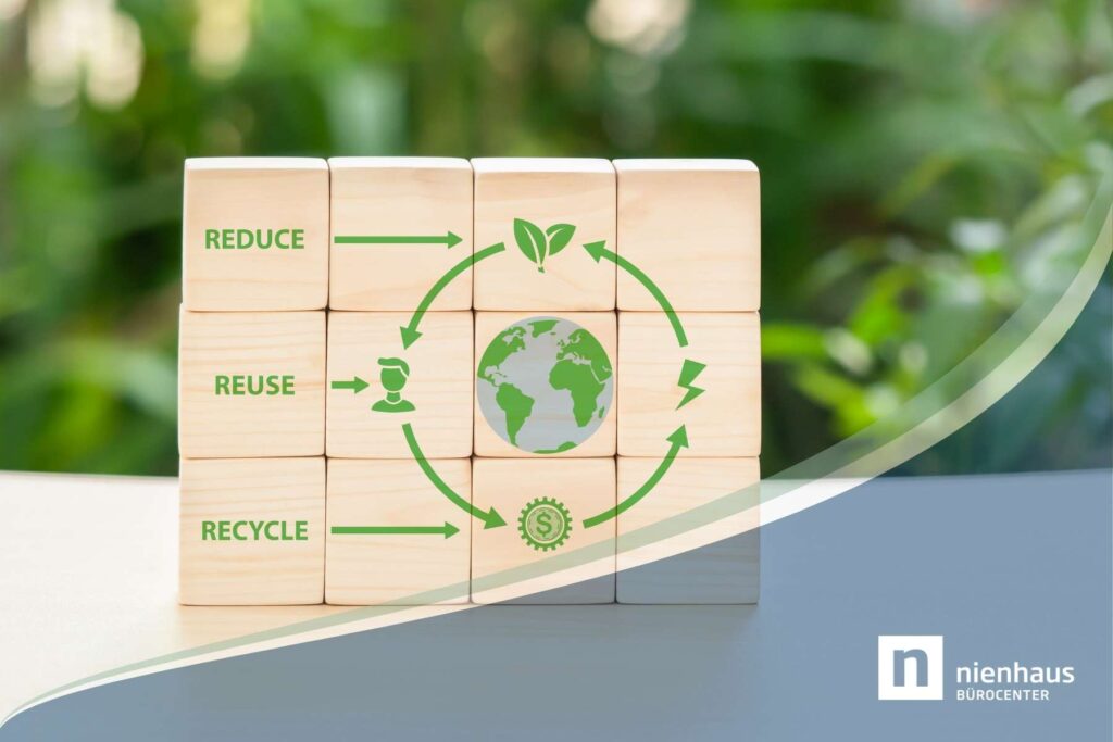Auf einem hellen Tisch stehen Bauklötze aus Holz, auf denen Reduce, Reuse und Recycle steht, während im Hintergrund unscharf Pflanzen zu sehen sind.