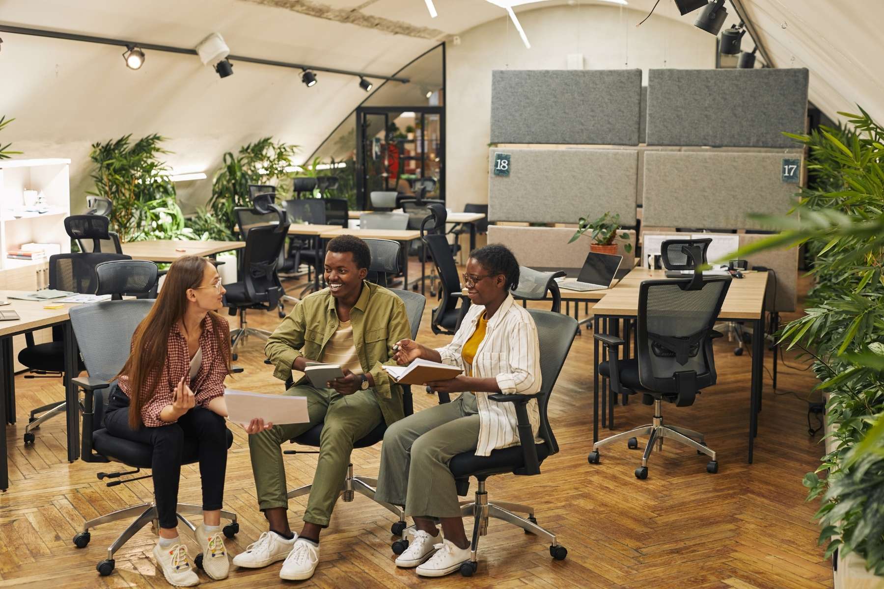Zwei weibliche und ein männlicher Angestellter sitzen lachend nebeneinander in einem freundlichen Büro, in dem viele Möbel aus Holz sind und viele Grünpflanzen aufgestellt wurden.