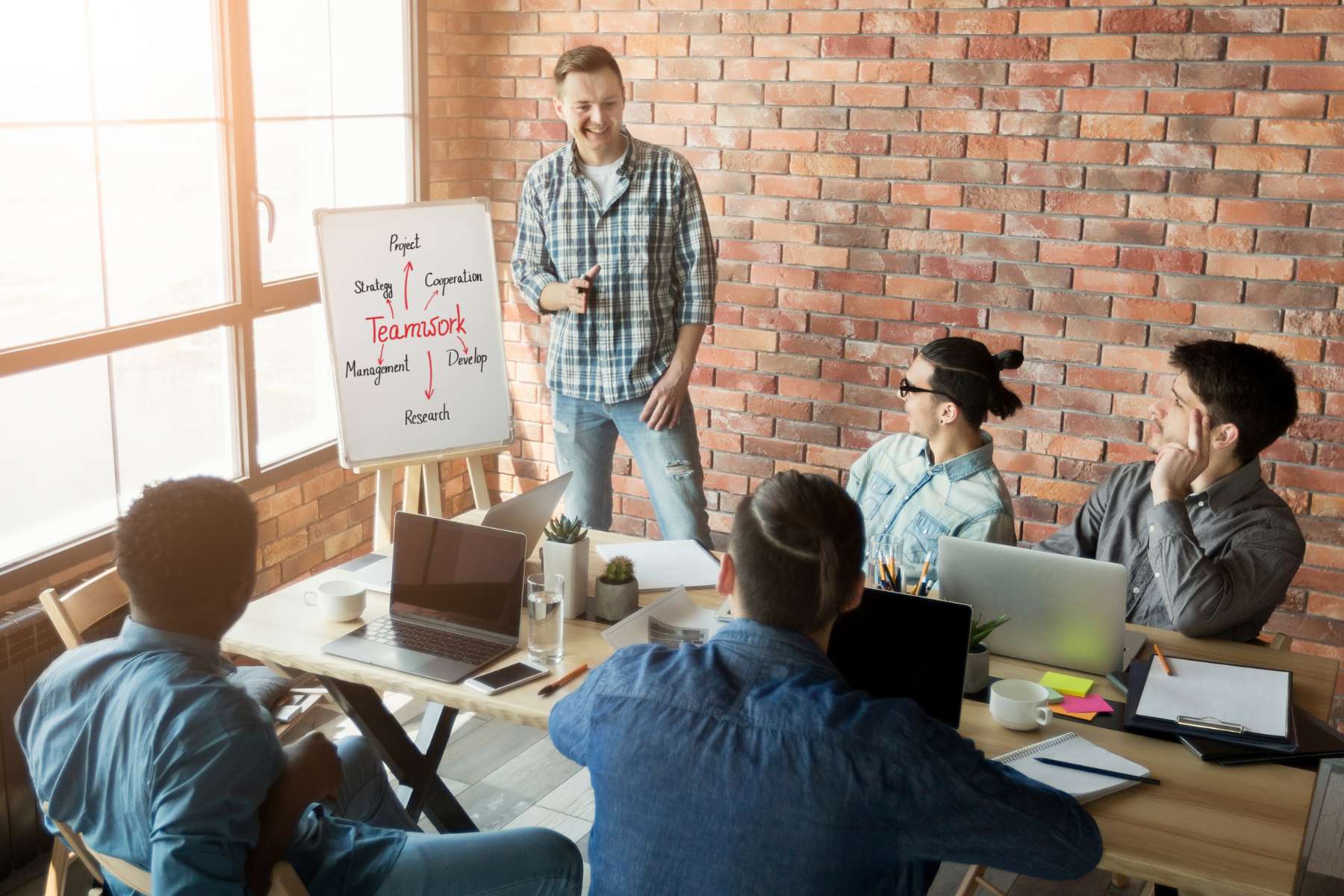 Ein männlicher Büroangestellter steht lächelnd neben einem Whiteboard, auf dem das Wort Teamwork erklärt wird, während vier weitere männliche Angestellte vor ihm an einem Tisch sitzen und zuhören.