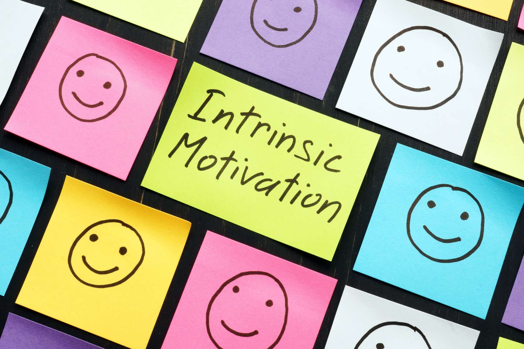 Eine Reihe bunter Post-Its nebeneinander und übereinander, auf denen je ein lächelndes Smiley abgebildet ist, während auf dem mittleren Post-It Intrinsic Motivation steht.