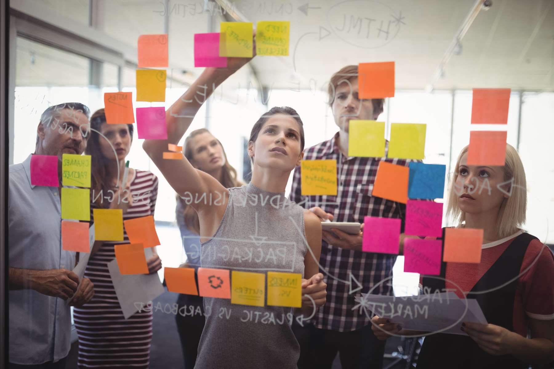 Eine Gruppe junger Angestellter schaut einer weiblichen Führungskraft konzentriert dabei zu, wie sie Post-Its an einer Glasscheibe anbringt.