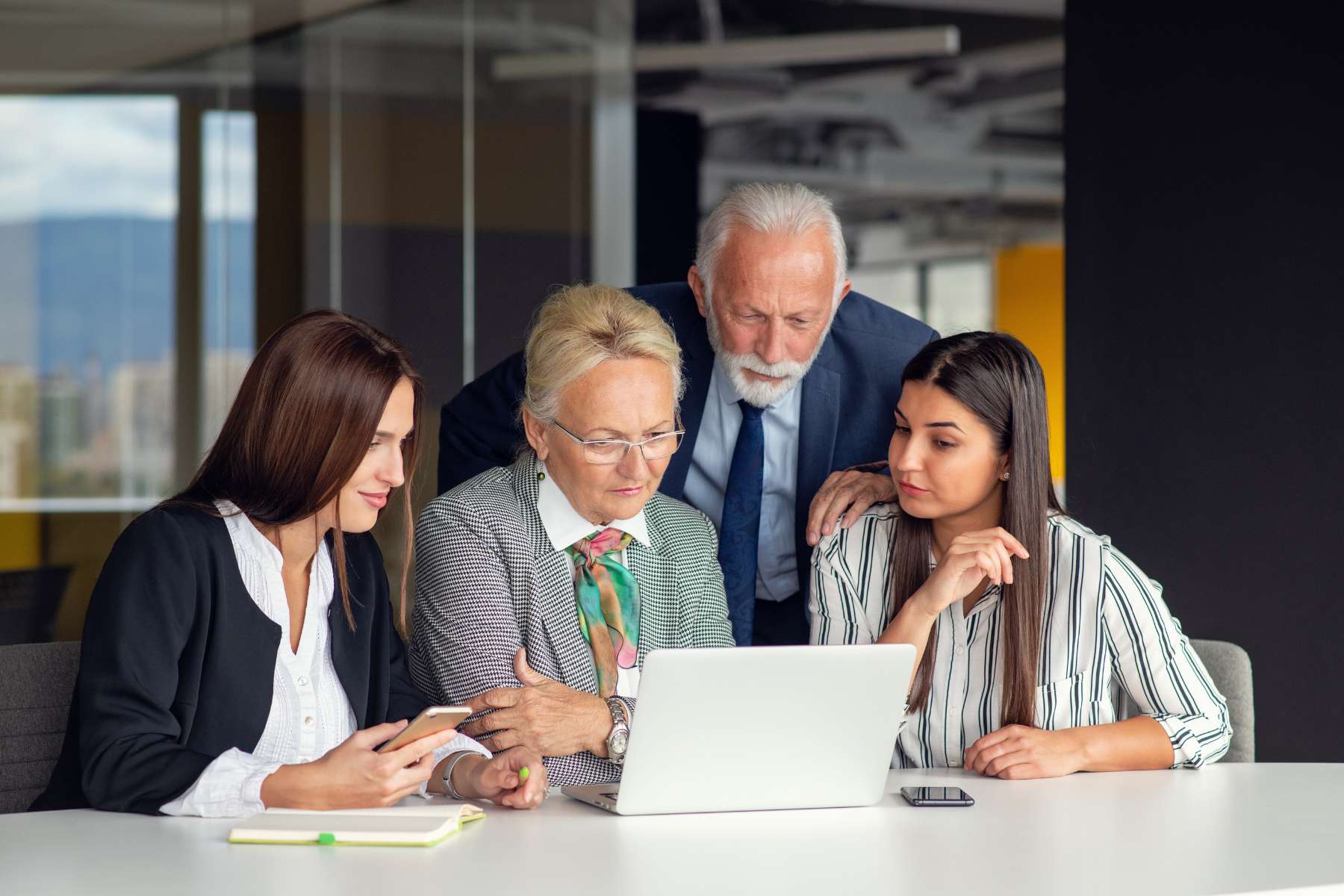 Zwei junge Mitarbeiterinnen aus der Generation Z sitzen mit einer wesentlich älteren Mitarbeiterin und einem ebenfalls wesentlich älteren Mitarbeiter zusammen im Büro am Tisch vor einem Laptop und schauen sich darauf etwas an.