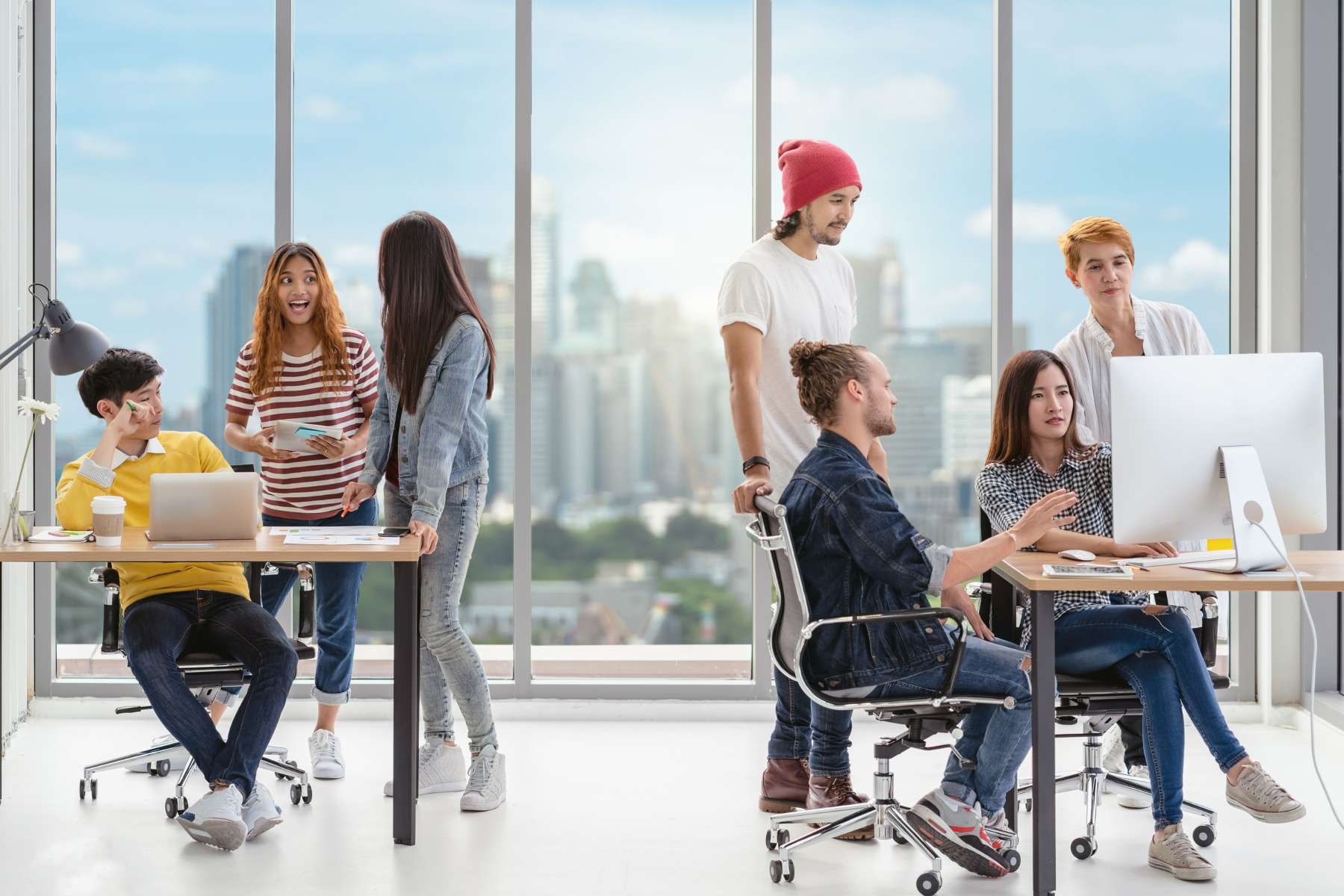 Eine Gruppe junger diverser Büroangestellter der Generation Z sitzt bzw. steht in zwei Gruppen um zwei Schreibtisch herum, arbeitet und unterhält sich.