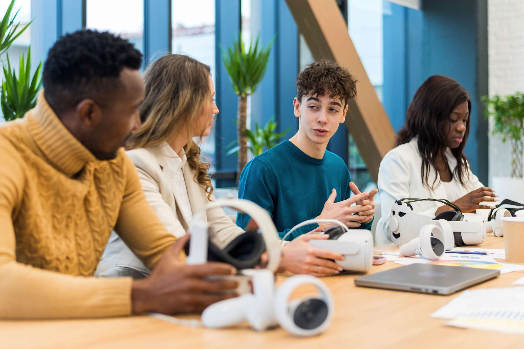 Zwei junge Männer und zwei junge Frauen der Generation Z sitzen an einem Holztisch, haben VR-Brillen vor sich liegen und unterhalten sich.
