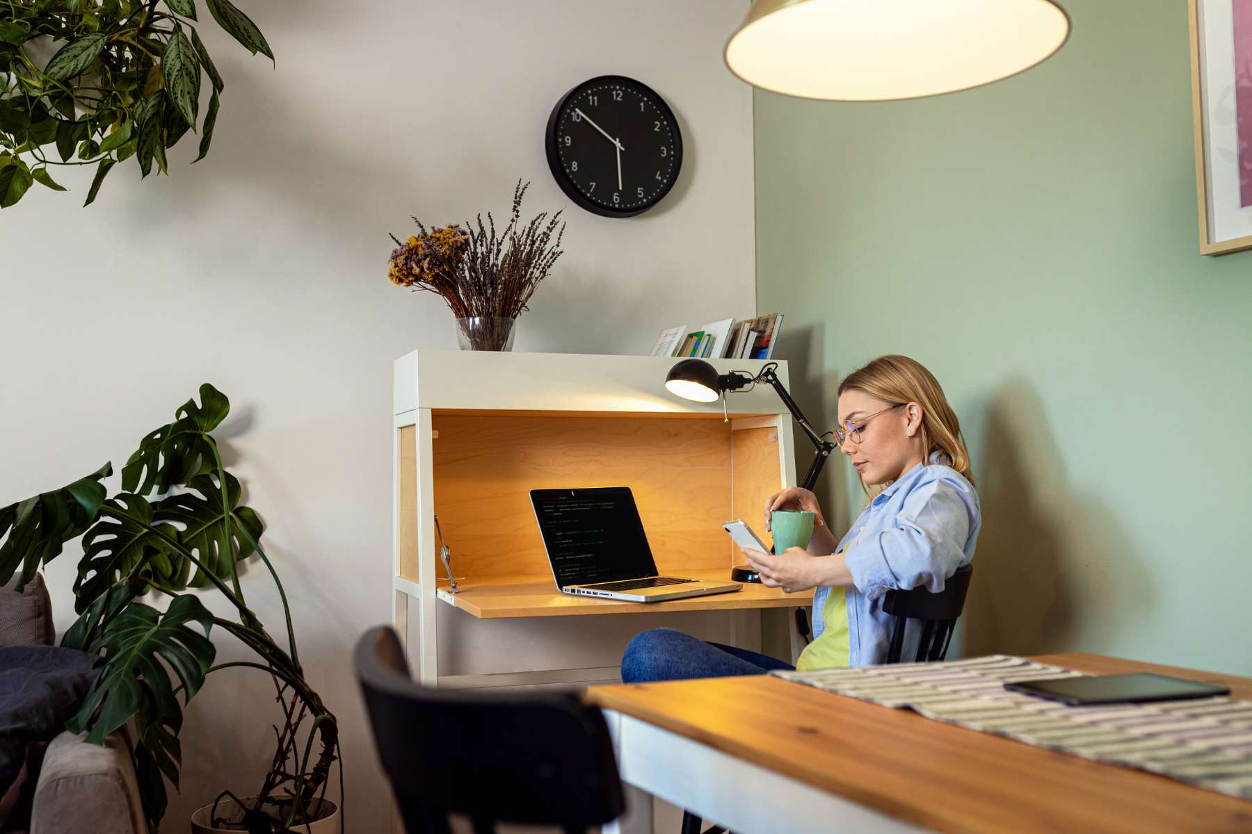 Junge Frau sitzt mit Handy und Kaffee in der Hand im Home-Office an einer Kommode mit ausklappbarer Tischplatte und arbeitet am Laptop.