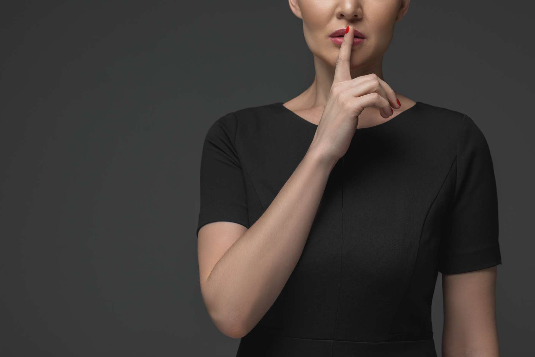 Eine Frau im schwarzen Shirt steht vor einem dunklen Hintergrund und hält sich als Geste des Schweigens den Zeigefinger vor die geschlossenen Lippen.