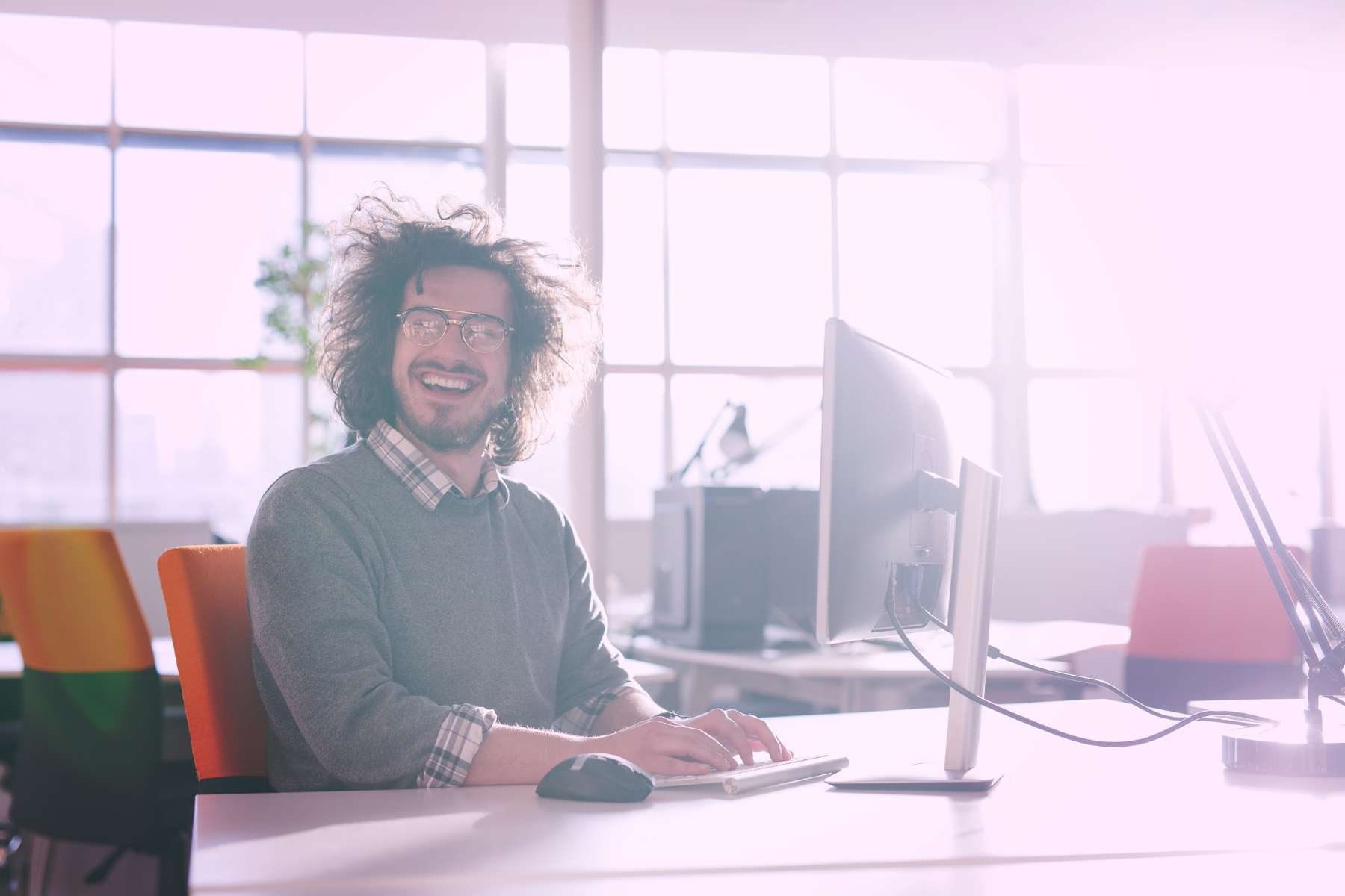 Ein glücklicher Mann mit Brille und zerzausten Haaren sitzt in einem hellen Büro am Schreibtisch und arbeitet am PC.