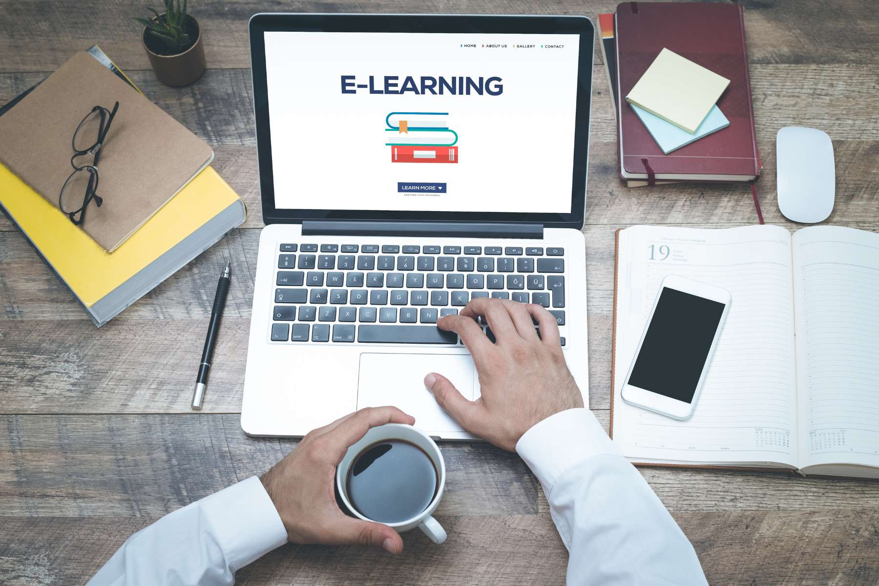 Aufnahme eines Holztisches aus der Vogelperspektive, auf dem Notizbücher, Stifte und ein Handy liegen und an dem ein Mann mit Kaffeetasse in der Hand an einem Laptop arbeitet und eine Session Microlearning im Büro macht.