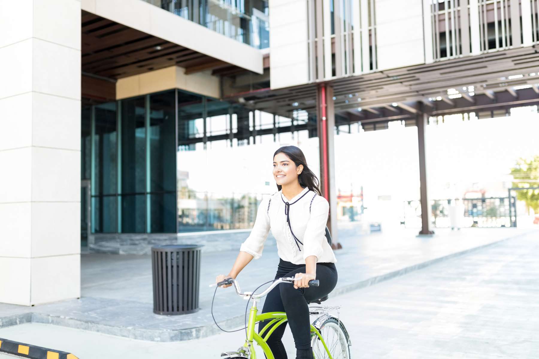 Eine junge Büroangestellte mit weißer Bluse und schwarzer Hose sitzt auf einem grünen Fahrrad und fährt weg vom Bürogebäude hinter ihr.