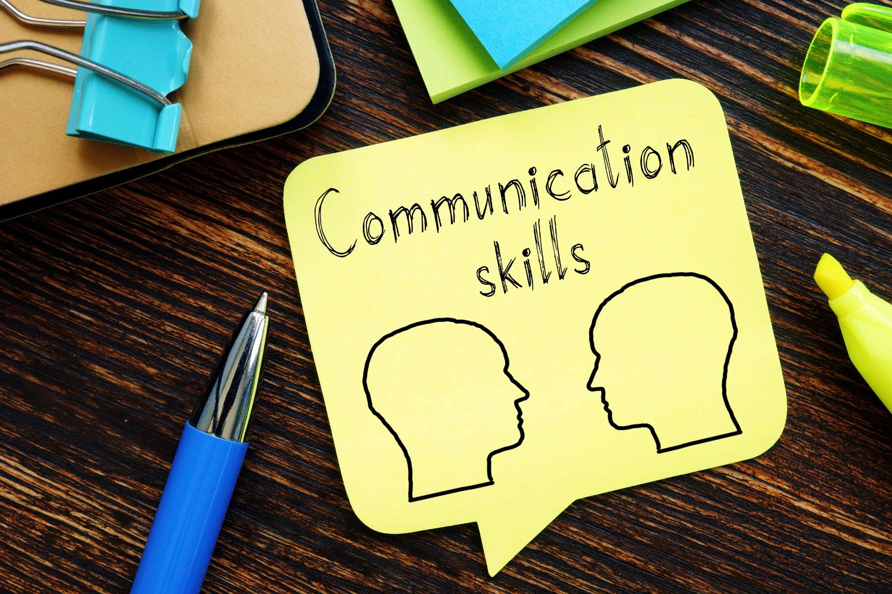 Neben Stiften liegt ein gelber Notizzettel mit 2 gezeichneten Köpfen und den Wörtern Communication Skills