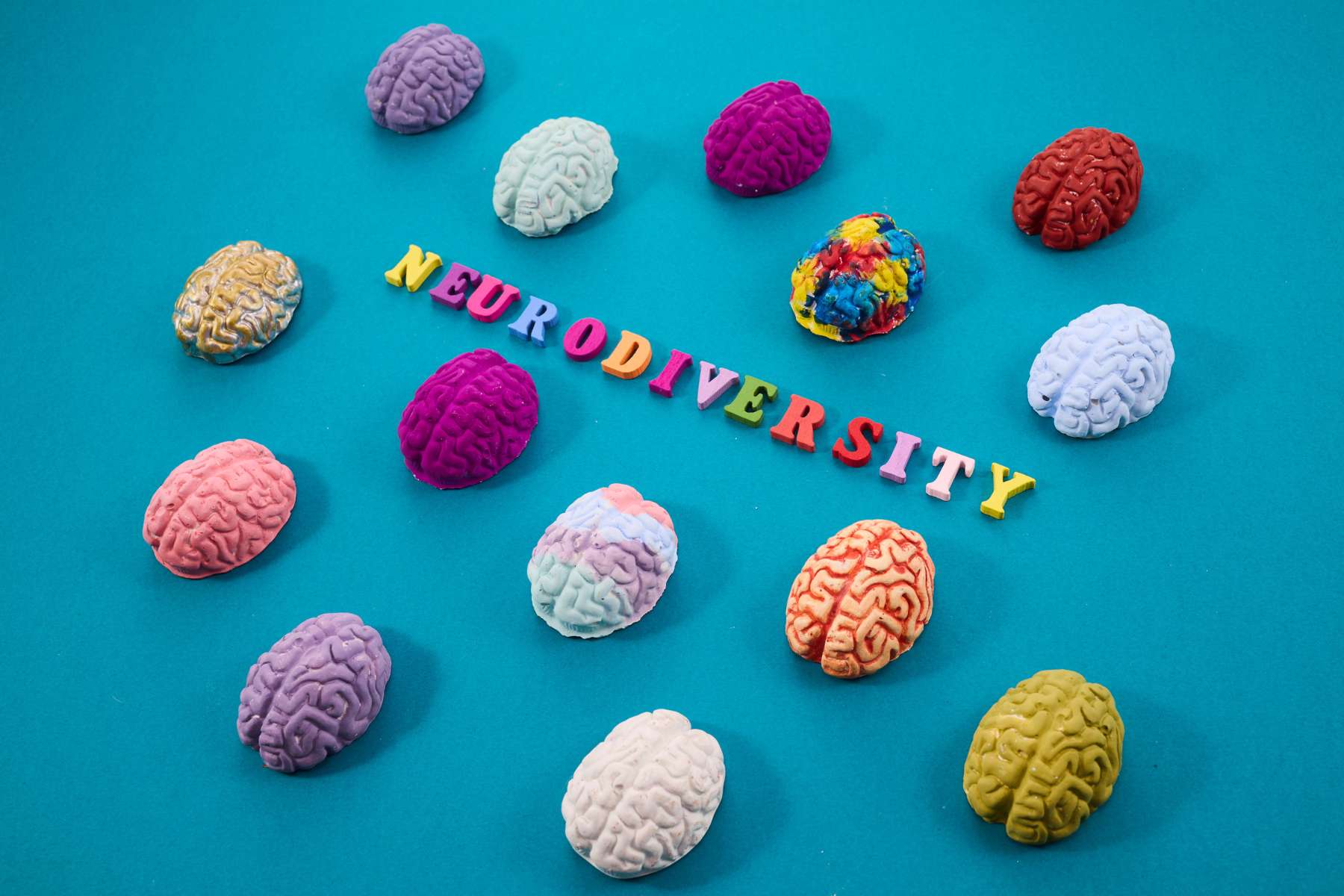 Auf einem türkisen Boden liegen verschiedenfarbige Nachstellungen von Gehirnen, als Sinnbild für neuroinklusive Arbeitswelten, während in der Mitte das Wort Neurodiversity in bunter Schrift steht.