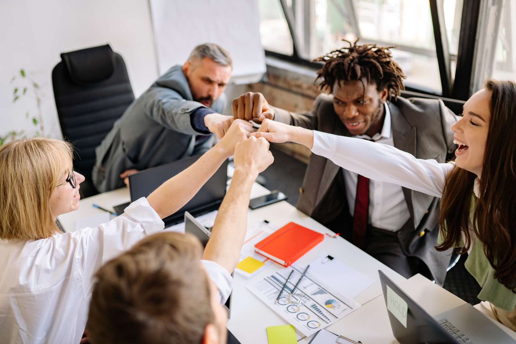 Ein 5-köpfiges Team sitzt im Büro, hält ein erfolgreiches Teammeeting ab und faustet sich in der Luft gegenseitig zu.